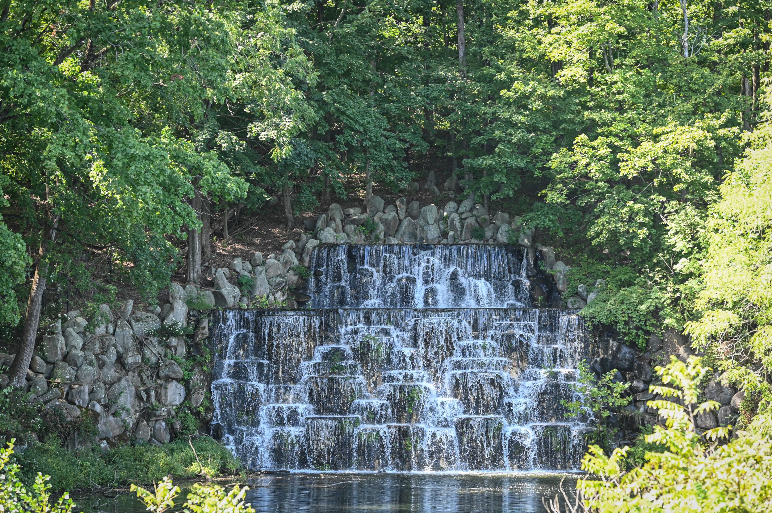 Image of a man-made cascading waterfall at Duke Farms, featuring water flowing over a series of rocky tiers surrounded by lush green foliage. The waterfall is structured in multiple levels, creating a picturesque scene, with sunlight filtering through the trees, enhancing the natural beauty of the setting.