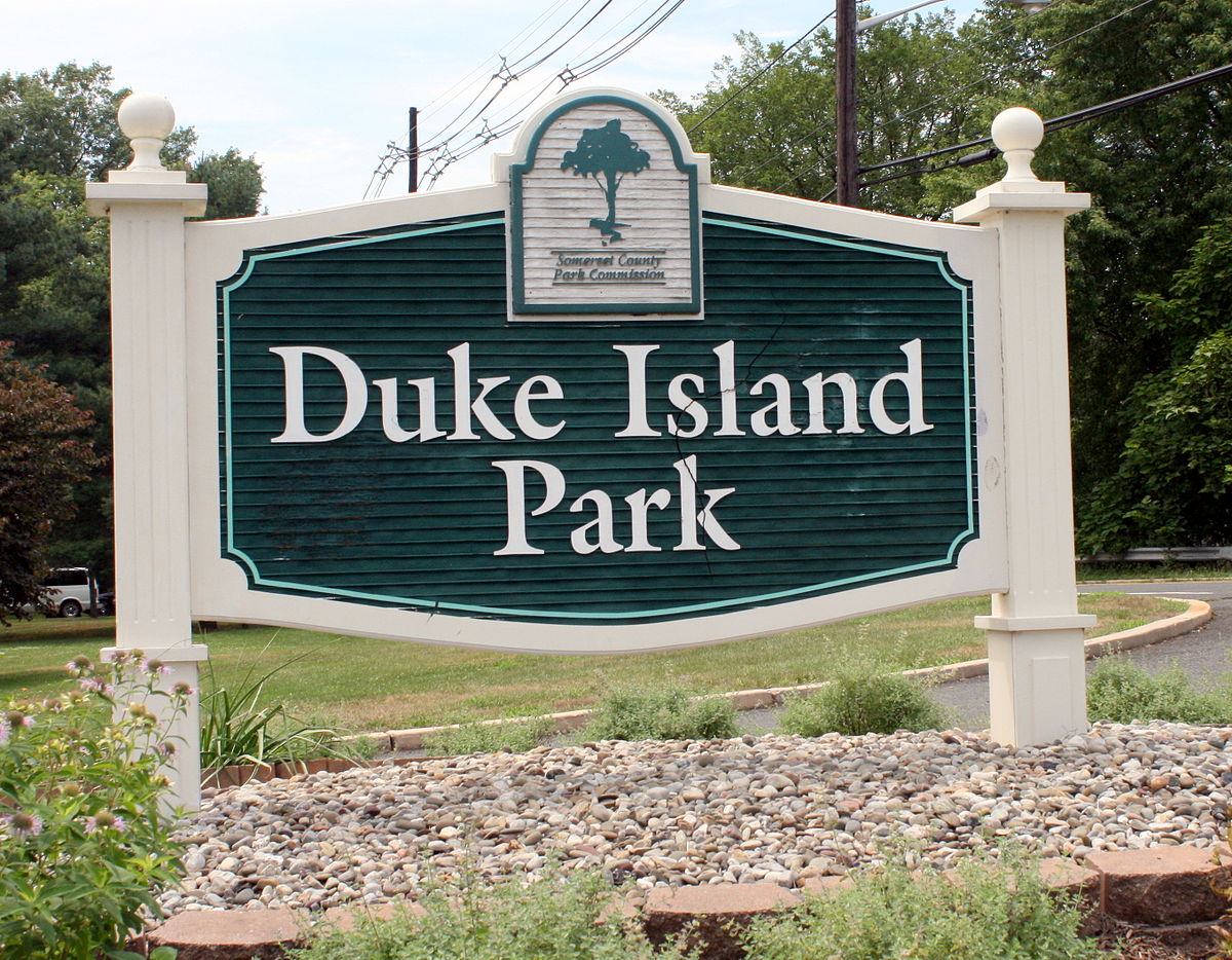 Signage for Duke Island Park, featuring a large dark green sign with bold, white lettering. The sign is supported by white columns and set against a backdrop of trees and a partly cloudy sky. A small logo with a tree and a description 'Somerset County Park Commission' is displayed at the top of the sign.