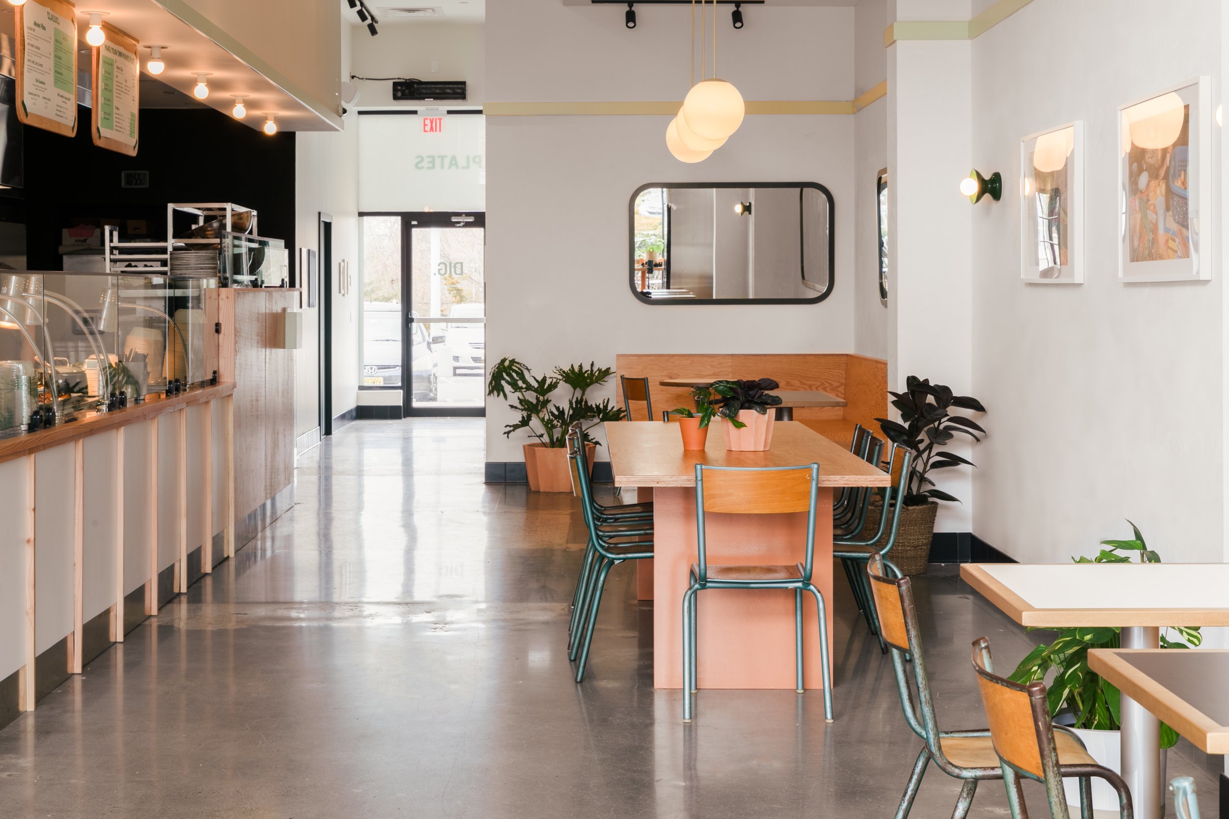 Interior view of DIG in Brindgewater NJ with a clean, minimalist design. The place features a combination of light wooden tables and orange metal chairs, with pendant lighting above and decorative plants adding a touch of greenery. A service counter with a glass display showcases various pastries, and large mirrors on the wall create a spacious feel. The entrance is visible in the background, with natural light streaming in.
